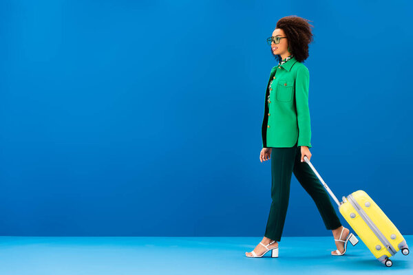 side view of smiling african american woman holding travel bag on blue background 