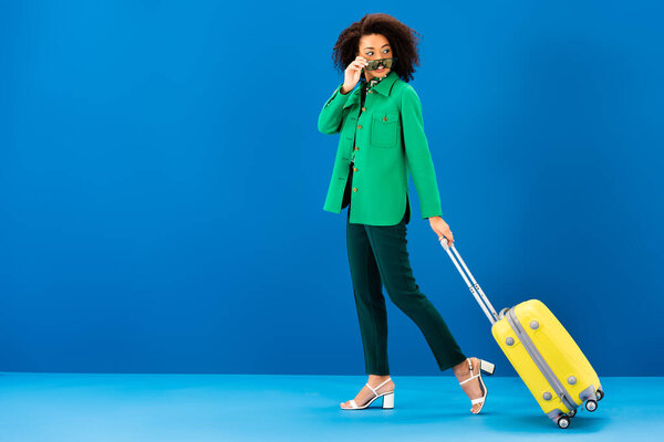smiling african american woman holding sunglasses and holding travel bag on blue background 