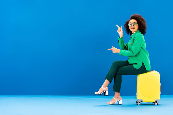 smiling african american woman pointing with fingers and sitting on travel bag on blue background 