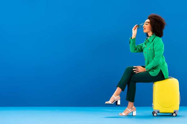 side view of smiling african american woman sitting on travel bag and putting on sunglasses on blue background 