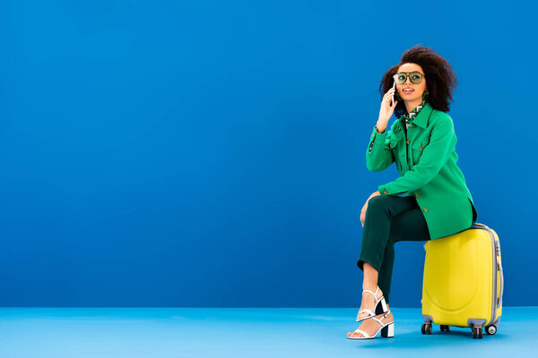 smiling african american woman sitting on travel bag and talking on telephone on blue background 