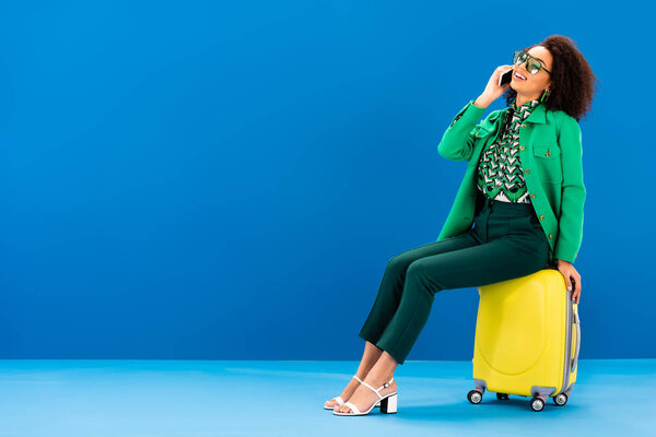 smiling african american woman sitting on travel bag and talking on smartphone on blue background 