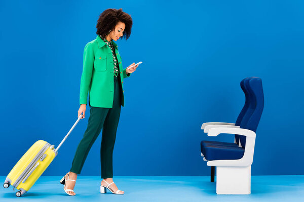 smiling african american woman holding travel bag and using smartphone on blue background 