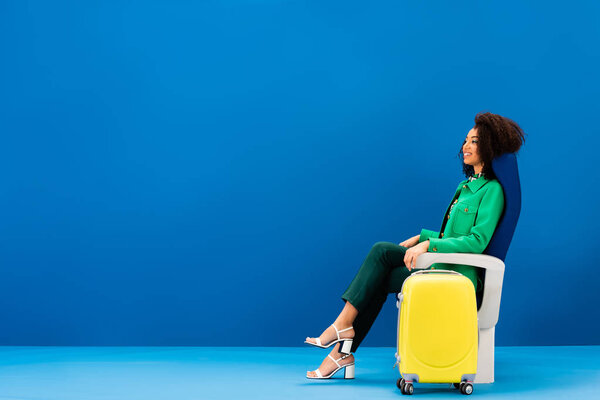 smiling african american woman sitting on seat near travel bag on blue background 