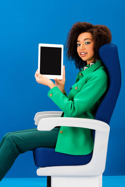 smiling african american sitting on seat and holding digital tablet on blue background 