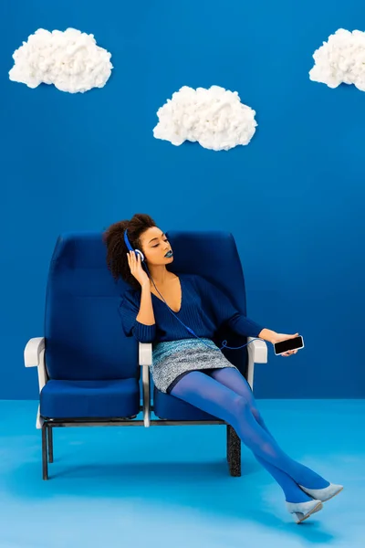 アフリカ系アメリカ人は席に座り音楽を聴き雲の中で青い背景にスマートフォンを持っています — ストック写真