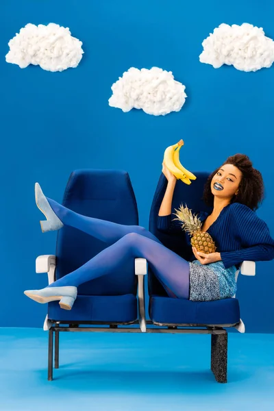 笑着的非洲裔美国人坐在座位上 拿着香蕉和菠萝 蓝蓝的背景上挂满了云彩 — 图库照片