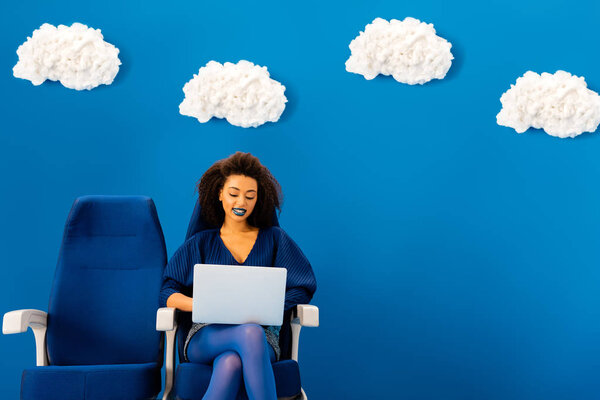 улыбающийся африканский американец сидит на сиденье и использует ноутбук на синем фоне с облаками
 