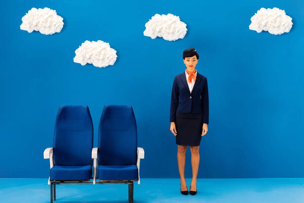 smiling african american flight attendant standing near seat on blue background with clouds 