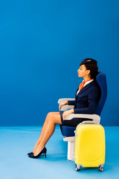 side view of african american flight attendant sitting on seat near travel bag on blue background 