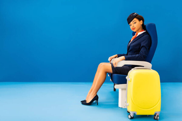 african american flight attendant sitting on seat near travel bag on blue background 