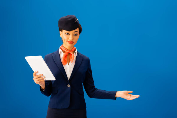 smiling african american flight attendant holding digital tablet and pointing with hand isolated on blue 