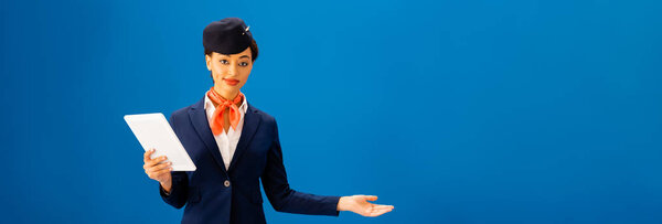 panoramic shot of smiling african american flight attendant holding digital tablet and pointing with hand isolated on blue 