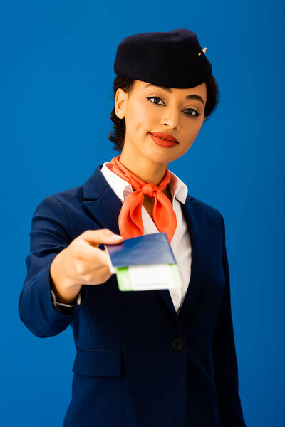 smiling african american flight attendant holding passport and air ticket isolated on blue 