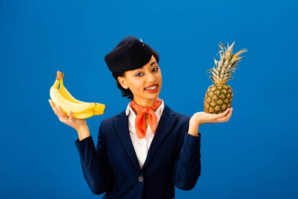 smiling african american flight attendant holding pineapple and bananas isolated on blue 
