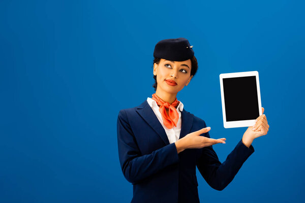 smiling african american flight attendant pointing with hand at digital tablet isolated on blue 