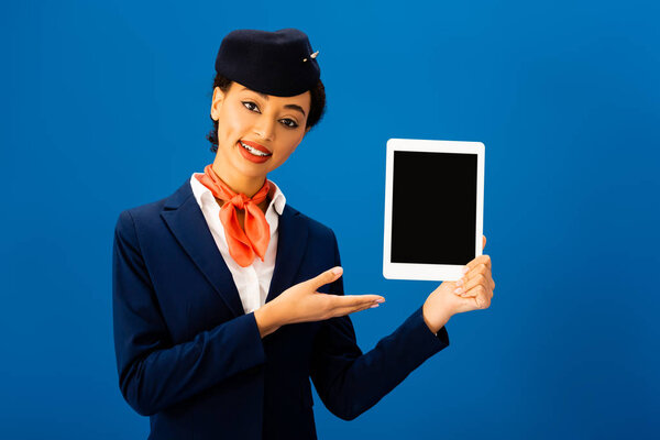 smiling african american flight attendant pointing with hand at digital tablet isolated on blue 