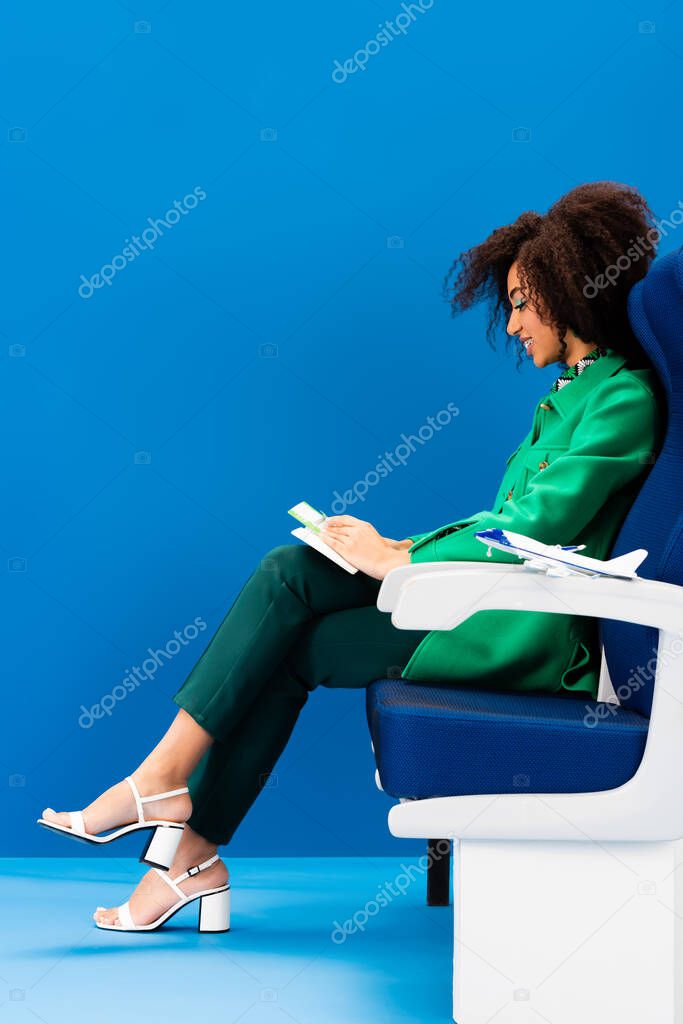 side view of smiling african american woman and model of plane on seat on blue background 