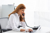 Seitenansicht einer lächelnden Geschäftsfrau, die am Telefon spricht und Laptop auf dem Tisch benutzt 