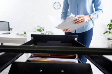 Cropped view of businesswoman holding folder with dossier near open cabinet driver in office