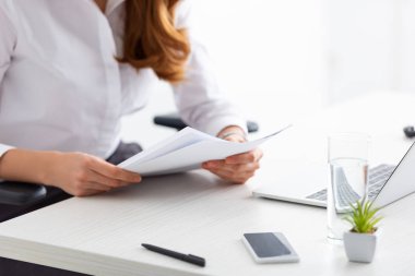 Cropped view of businesswoman holding papers with dossier at table with laptop and smartphone