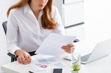 Cropped view of businesswoman holding paper with graphs near smartphone and laptop on table