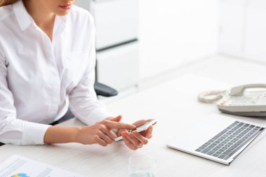 Cropped view of businesswoman using smartphone near document with chart and laptop on table