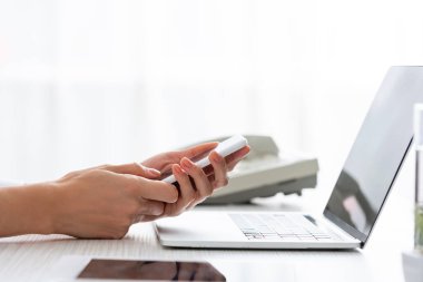 Cropped view of businesswoman using smartphone near digital tablet and laptop on table