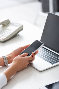 Cropped view of businesswoman using smartphone near laptop and digital tablet on table