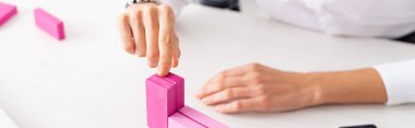 Cropped view of businesswoman stacking pink building blocks on table, panoramic shot