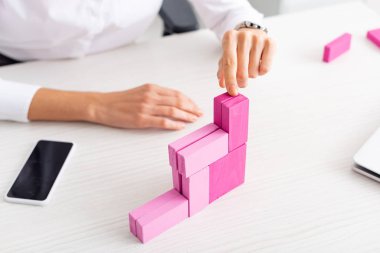 Cropped view of businesswoman stacking marketing pyramid from pink building blocks near gadgets on table