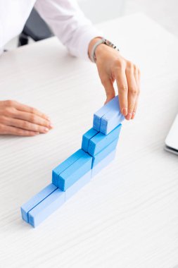Cropped view of businesswoman stacking blue building blocks near laptop on table