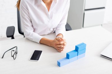 Cropped view of businesswoman sitting at table with stacked blue building blocks and gadgets