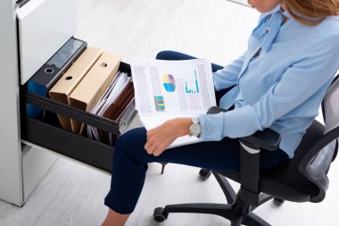 High angle view of  businesswoman holding papers with charts while sitting near open cabinet driver