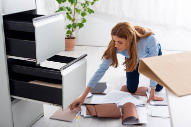 Businesswoman searching dossier near documents and contract on floor in office
