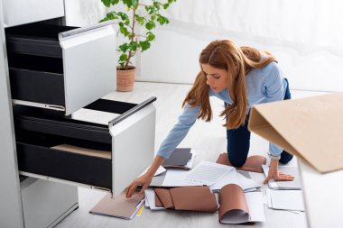 Businesswoman searching dossier near documents and contract on floor in office clipart