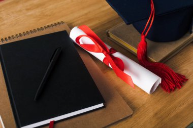 Not defterleri, kalem, diploma ve tahta arka planda mezuniyet şapkası.