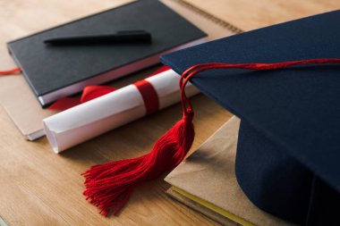 Tahta zemin üzerinde kalem, diploma ve mezuniyet şapkası bulunan defterlerin seçici odak noktası