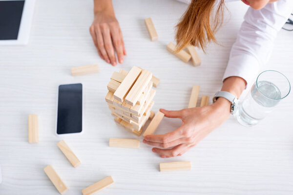 High angle view of businesswoman stacking building blocks near gadgets and glass of water on table 
