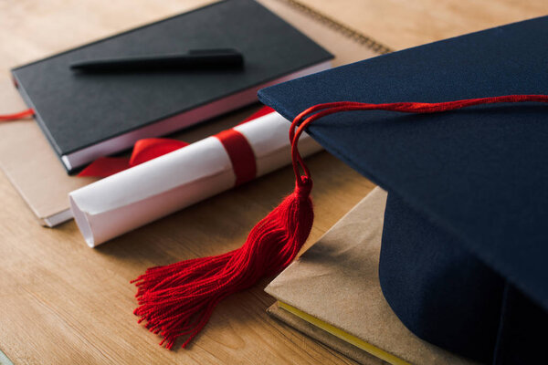 Selective focus of notebooks with pen, diploma and graduation cap on wooden background