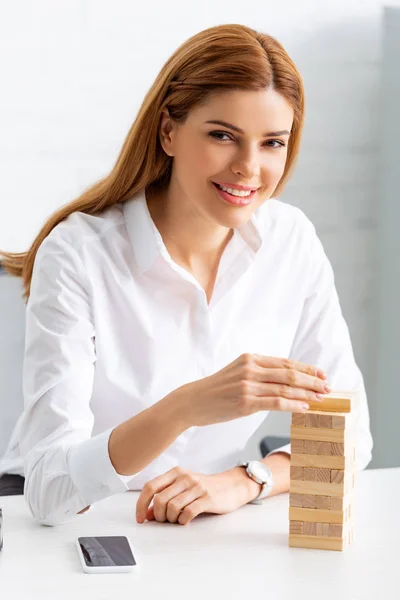 Smiling businesswoman looking at camera and stacking wooden building blocks on table — Stock Image Smiling Businesswoman Looking Camera Stacking Wooden Building Blocks Table Stock Photo