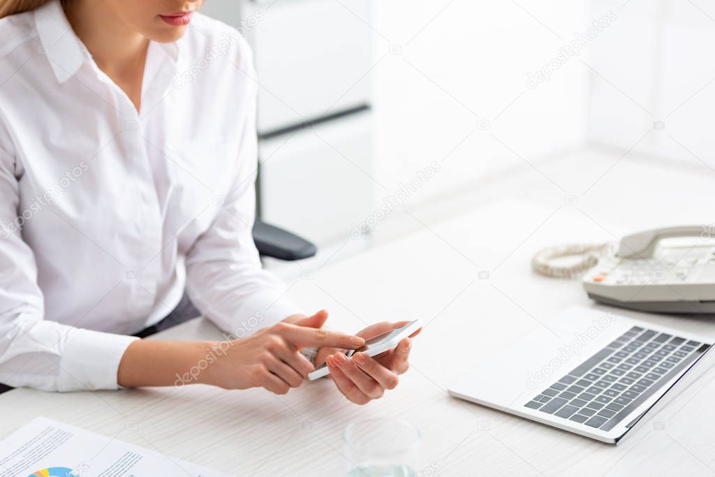 Cropped view of businesswoman using smartphone near document with chart and laptop on table