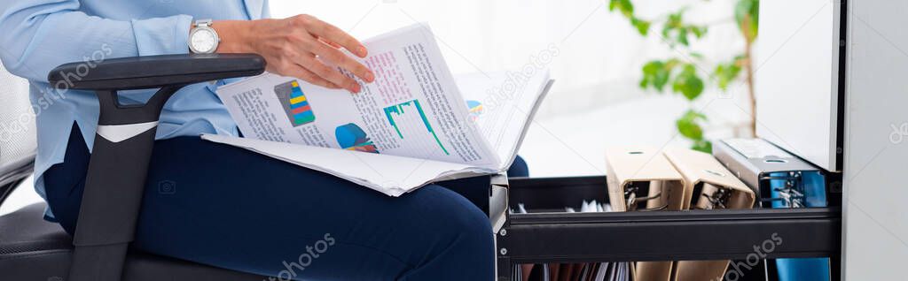 Cropped view of businesswoman holding papers with graphs while sitting near open cabinet driver, panoramic shot 