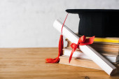 Diplom s krásným lukem a maturitní čepice s červeným střapcem na vrcholu knihy na stole na bílém pozadí