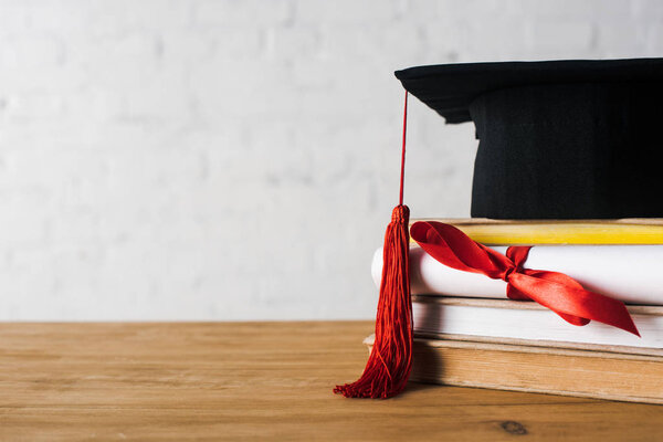 Diploma with beautiful bow, graduation cap with red tassel on top of books on table on white background