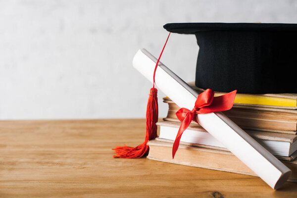 Diploma with beautiful bow and graduation cap with red tassel on top of books on table on white background