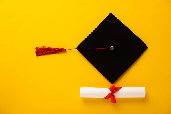 Top view of diploma with beautiful bow and graduation cap with red tassel on yellow background