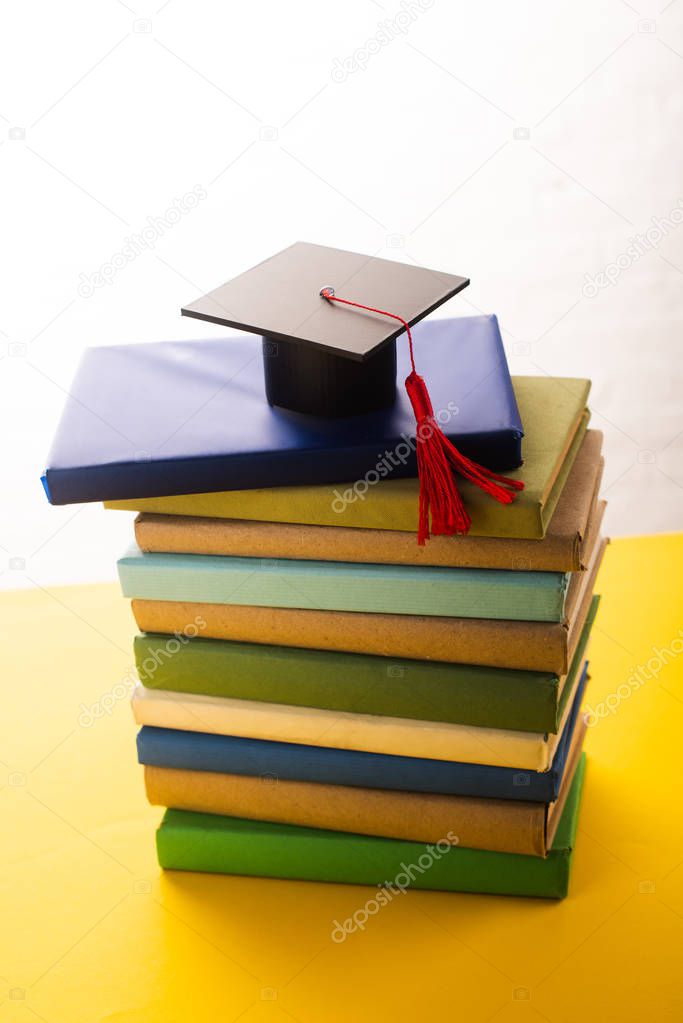 Graduation cap with red tassel on top of books on yellow surface on white background