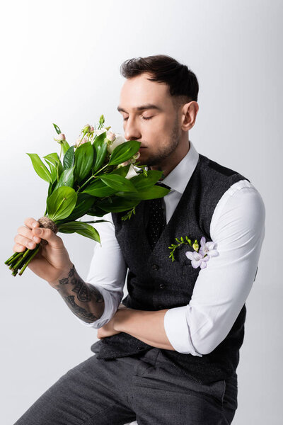 handsome tattooed elegant bridegroom smelling bouquet with closed eyes on grey