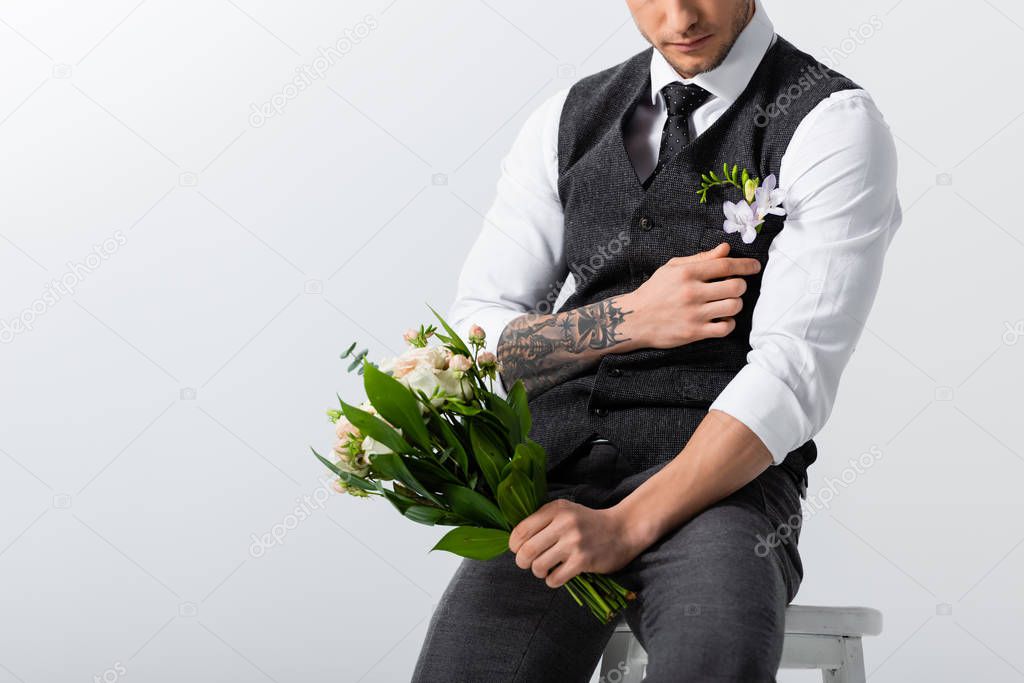 cropped view of tattooed elegant bridegroom with bouquet adjusting boutonniere on grey